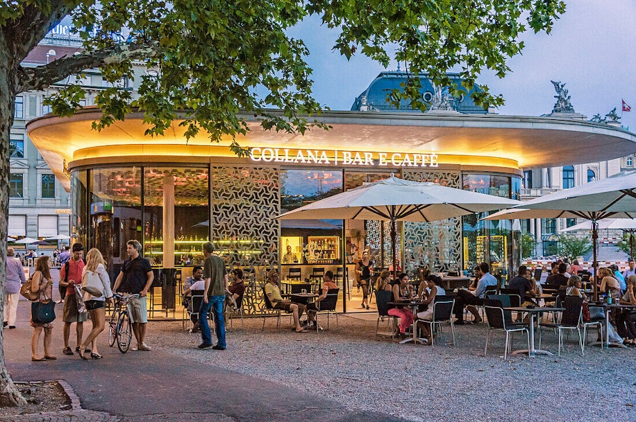 Ein modernes, rundes Café mit Glaswänden erstrahlt abends in warmem Licht, wenn Menschen an den Tischen im Freien sitzen und andere in der Nähe spazieren oder stehen. Sonnenschirme spenden Schatten, und grüne Bäume rahmen das lebhafte Ambiente ein.