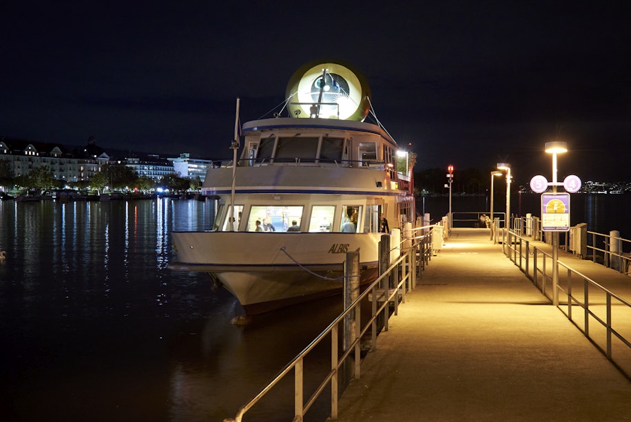Ein weißes Boot namens Albis liegt nachts an einem Pier. Warmes Licht erhellt den Steg und spiegelt sich im ruhigen Wasser. Im Hintergrund sind Gebäude und Lichter auf der anderen Seite des Wassers zu sehen.