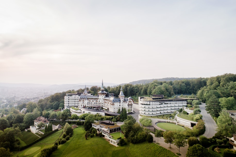 Luftaufnahme eines großen, eleganten Hotelkomplexes mit weißen Gebäuden, umgeben von üppigen grünen Bäumen und Rasenflächen, vor einer Kulisse aus sanften Hügeln und einem dunstigen Himmel.