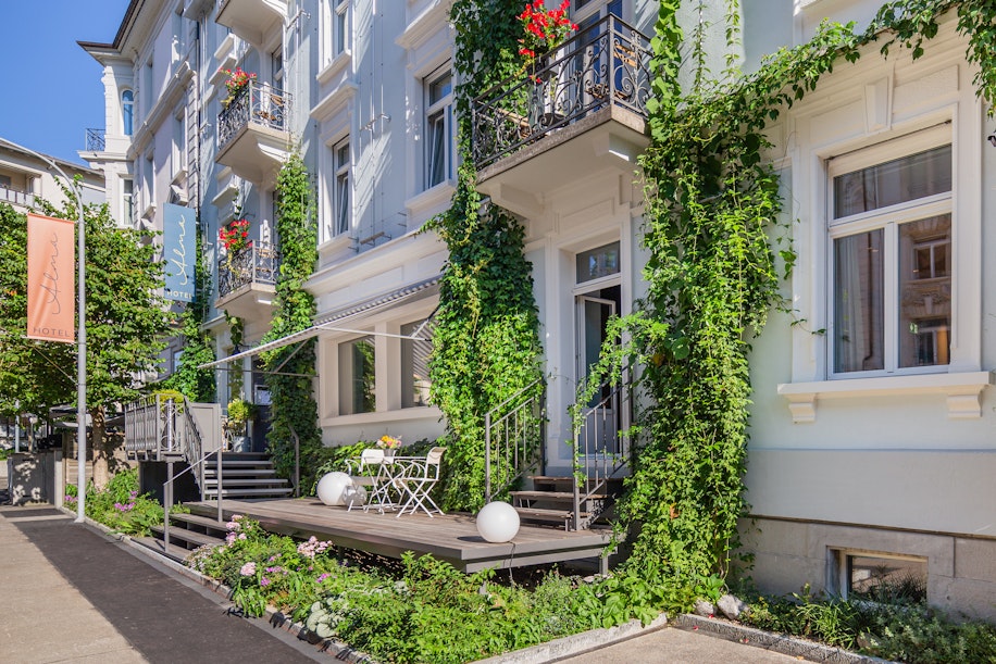Ein charmantes weißes Hotelgebäude mit Efeu an der Fassade, Balkonen mit roten Blumen und einem kleinen Außenbereich mit Tisch und Stühlen auf einer erhöhten Terrasse. Ein Hotelschild hängt auf der linken Seite am Bürgersteig.