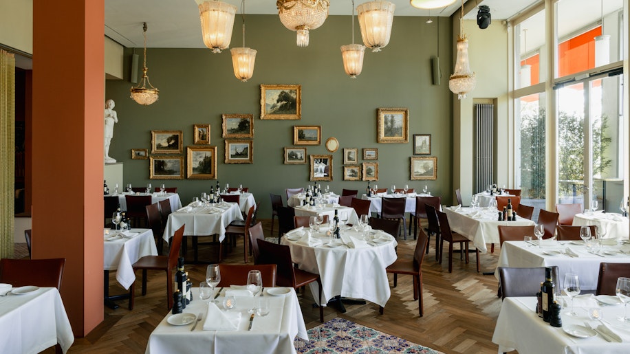 Eleganter Speisesaal des Restaurants mit runden, weiß gedeckten Tischen, Holzstühlen, Kronleuchtern, gerahmter Kunst an salbeigrünen Wänden und großen Fenstern, die viel Tageslicht hereinlassen.