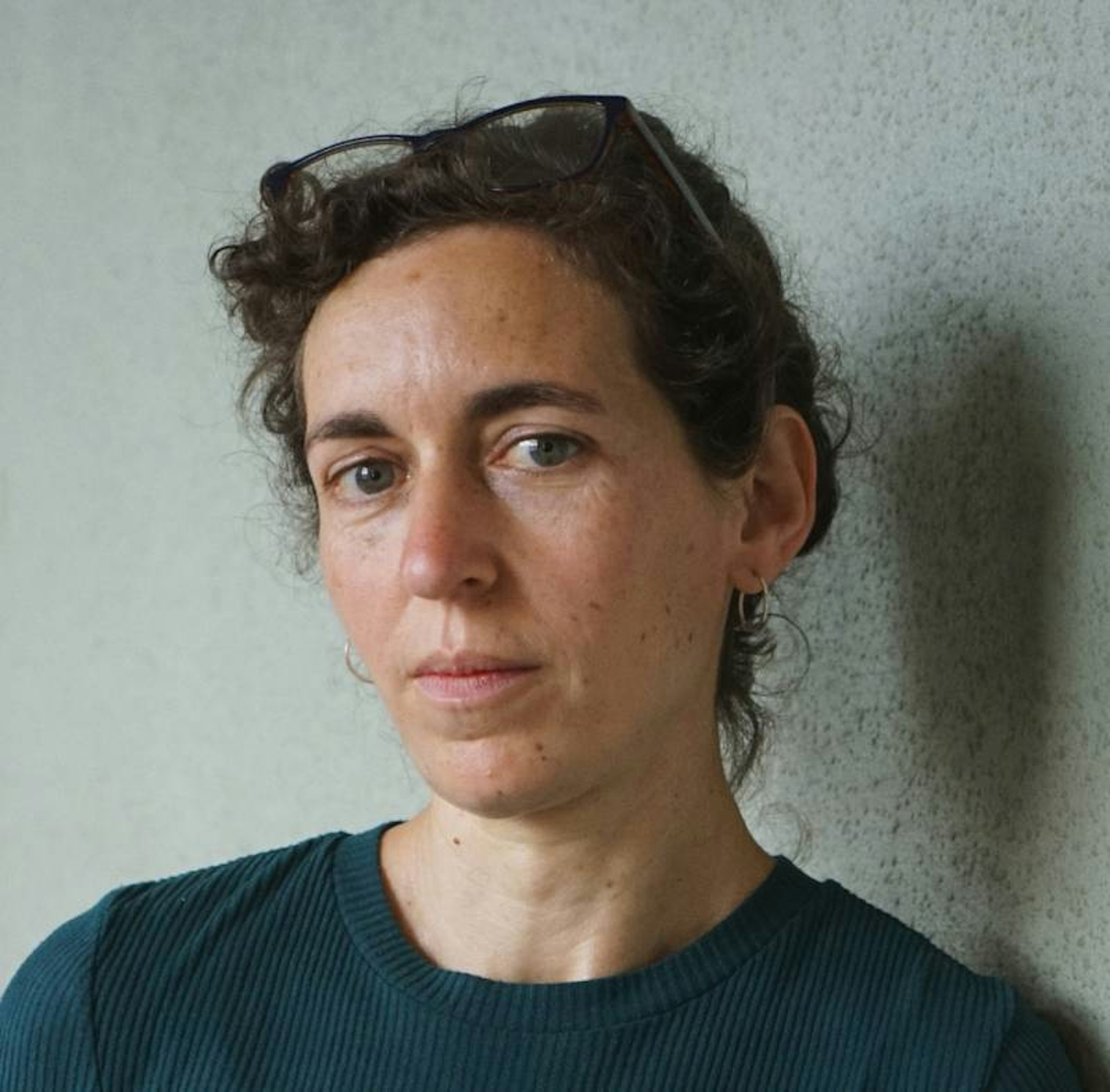 Eine Frau mit kurzen, lockigen braunen Haaren, die ein dunkelgrünes, geripptes Hemd trägt und eine Brille auf dem Kopf hat, steht an einer hellen, strukturierten Wand und schaut ruhig in die Kamera.