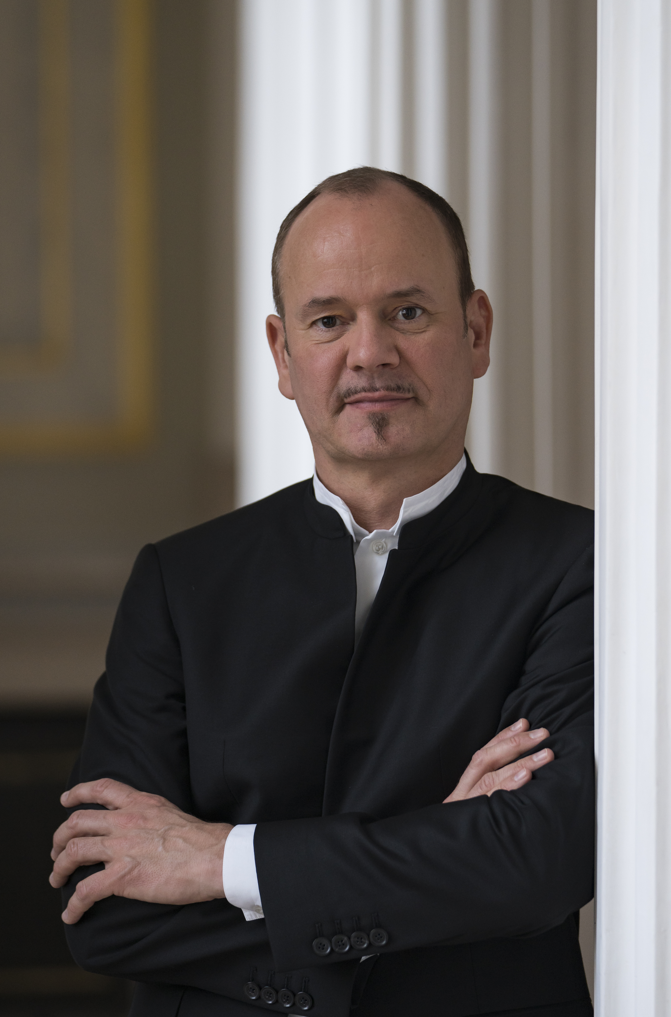 Middle-aged man in black suit, with crossed arms, leaning against doorway.