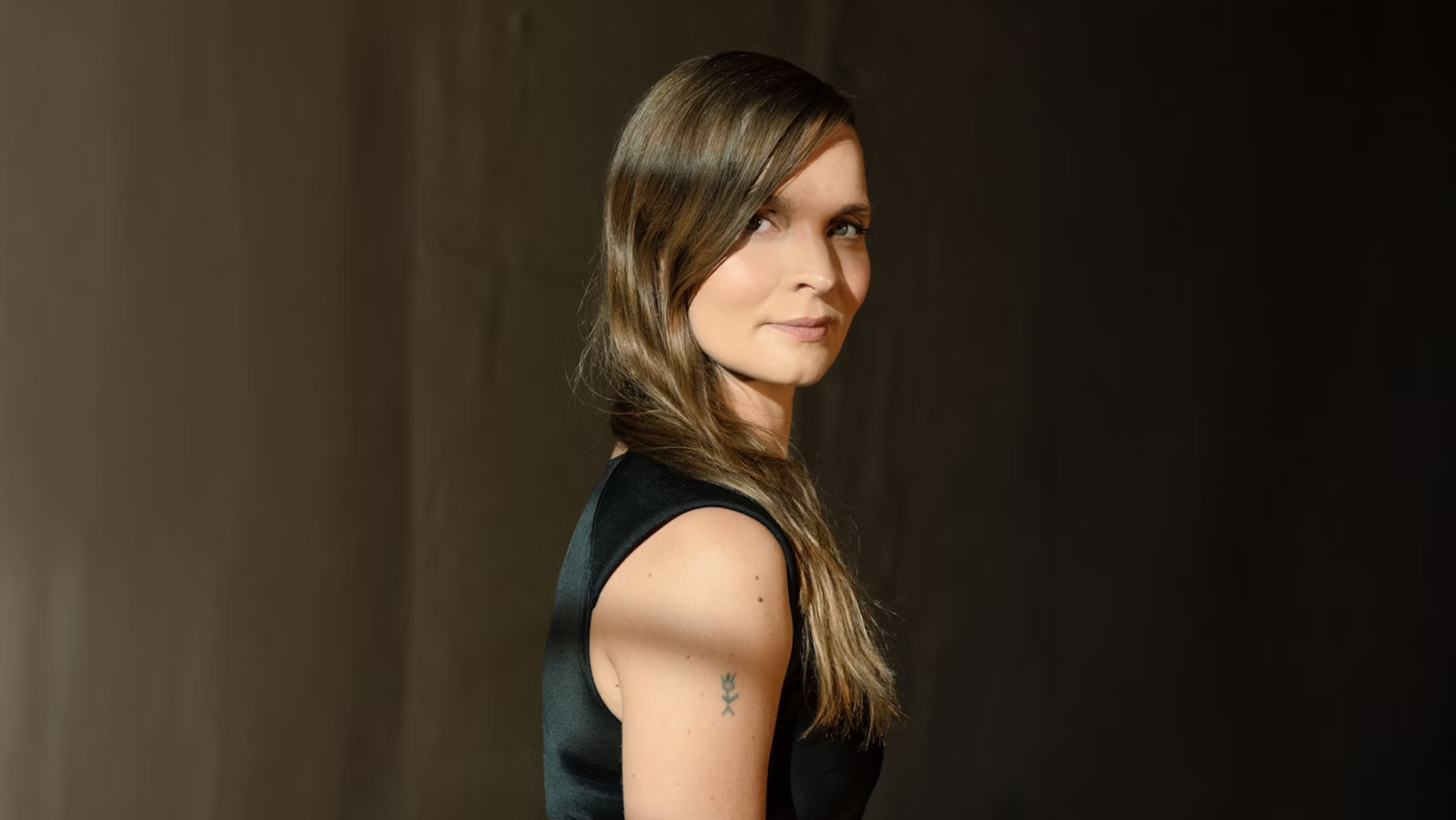 Eine Frau mit langen braunen Haaren, die ein ärmelloses schwarzes Oberteil trägt, steht im Profil vor einem dunklen Hintergrund. Das Licht hebt eine Seite ihres Gesichts und ihrer Haare hervor, und auf ihrem Oberarm ist eine kleine Tätowierung zu erkennen.
