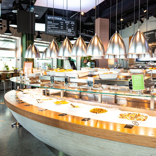 Ein modernes Buffet mit Wärmelampen aus Metall, Serviertabletts mit verschiedenen warmen und kalten Gerichten und einem geschwungenen Tresen in einem hell erleuchteten Restaurant.