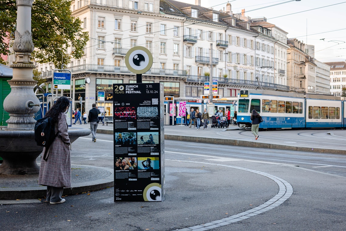 Eine Frau in einem langen Mantel steht in der Nähe einer Infotafel des Zurich Film Festival auf einer Straße in der Stadt, während eine Straßenbahn vorbeifährt und Menschen im Hintergrund spazieren gehen. Historische Gebäude säumen die Straße unter einem bewölkten Himmel.