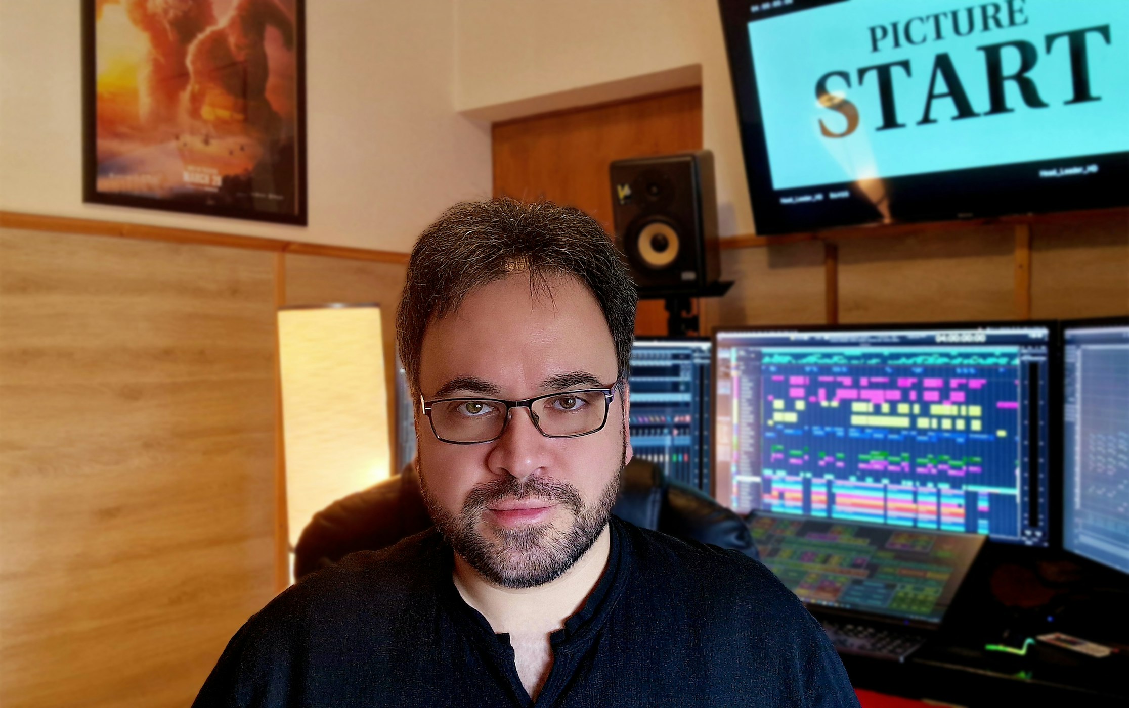Ein Mann mit Brille und dunklem Haar sitzt in einem Musikstudio, umgeben von Computermonitoren, die bunte Tonspuren anzeigen, Lautsprechern, einer Lampe und einem Bildschirm mit der Aufschrift PICTURE START. Ein Filmposter hängt an der Wand.