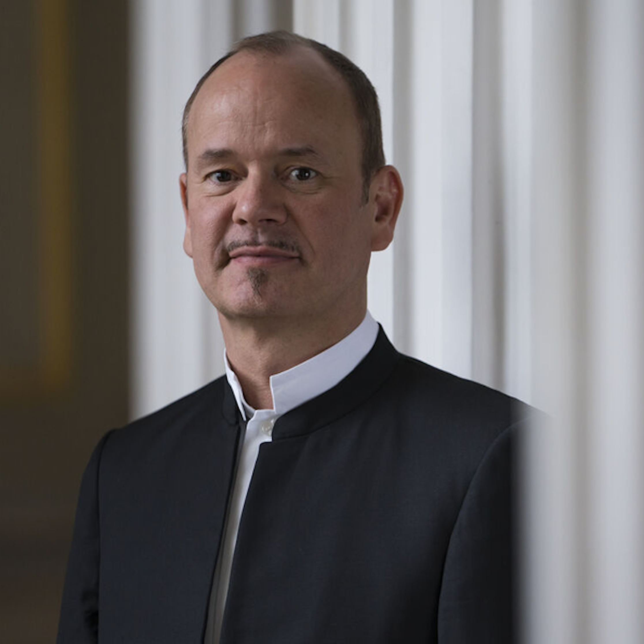 Ein Mann mit Schnurrbart und kurzen Haaren, der einen schwarzen Anzug und ein weißes kragenloses Hemd trägt, steht in einem Innenraum vor einer weißen Wand und blickt ruhig in die Kamera.