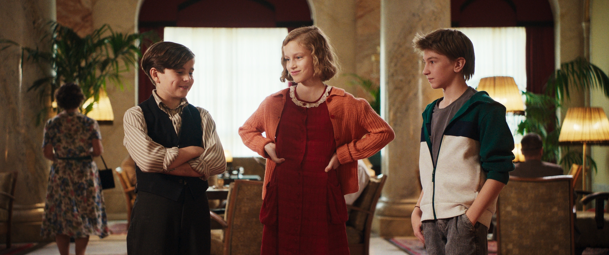 Drei Kinder stehen sich in einem Raum gegenüber, zwei Jungen und ein Mädchen in der Mitte. Das Mädchen trägt ein rotes Kleid und eine orangefarbene Strickjacke, die Jungen tragen Freizeitkleidung. Sie scheinen sich in einem warm beleuchteten, eleganten Raum mit Pflanzen zu unterhalten.