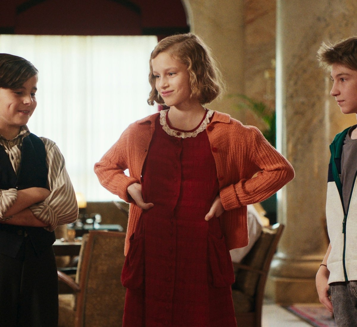 Das Geheime Stockwerk Film Still Drei Kinder stehen sich in einem Raum gegenüber, zwei Jungen und ein Mädchen in der Mitte. Das Mädchen trägt ein rotes Kleid und eine orangefarbene Strickjacke, die Jungen tragen Freizeitkleidung. Sie scheinen sich in einem warm beleuchteten, eleganten Raum mit Pflanzen zu unterhalten.