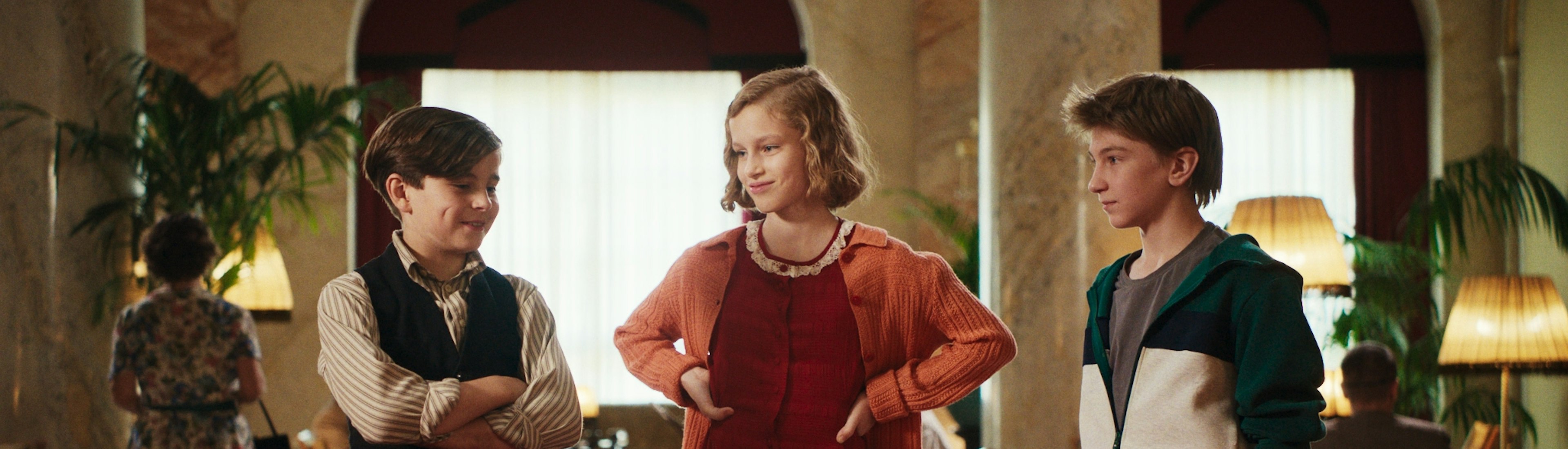 Das Geheime Stockwerk Film Still Drei Kinder stehen sich in einem Raum gegenüber, zwei Jungen und ein Mädchen in der Mitte. Das Mädchen trägt ein rotes Kleid und eine orangefarbene Strickjacke, die Jungen tragen Freizeitkleidung. Sie scheinen sich in einem warm beleuchteten, eleganten Raum mit Pflanzen zu unterhalten.