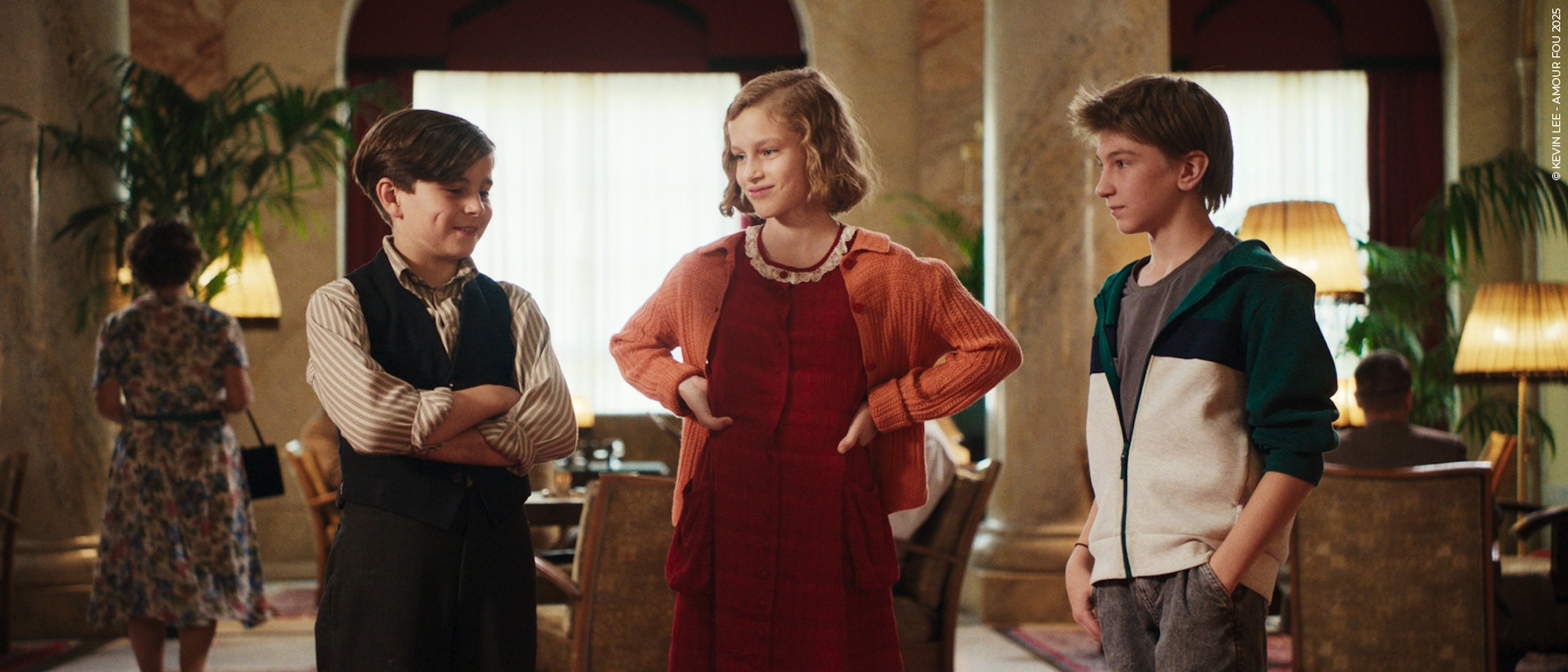 Drei Kinder stehen in einem schicken Raum mit Marmorsäulen und eleganten Lampen. Das Mädchen in der Mitte trägt ein rotes Kleid und eine Strickjacke, während die beiden Jungen links und rechts von ihr Freizeitkleidung tragen. Sie scheinen alle zu lächeln.