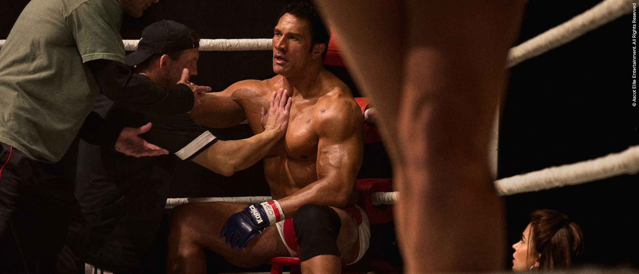 Ein muskulöser männlicher MMA-Kämpfer sitzt auf einem Hocker in der Ecke eines Boxrings und erhält zwischen den Runden Ratschläge und Unterstützung von seinen Trainern. Ein anderer Kämpfer steht im Vordergrund, eine Frau hockt an den Seilen.