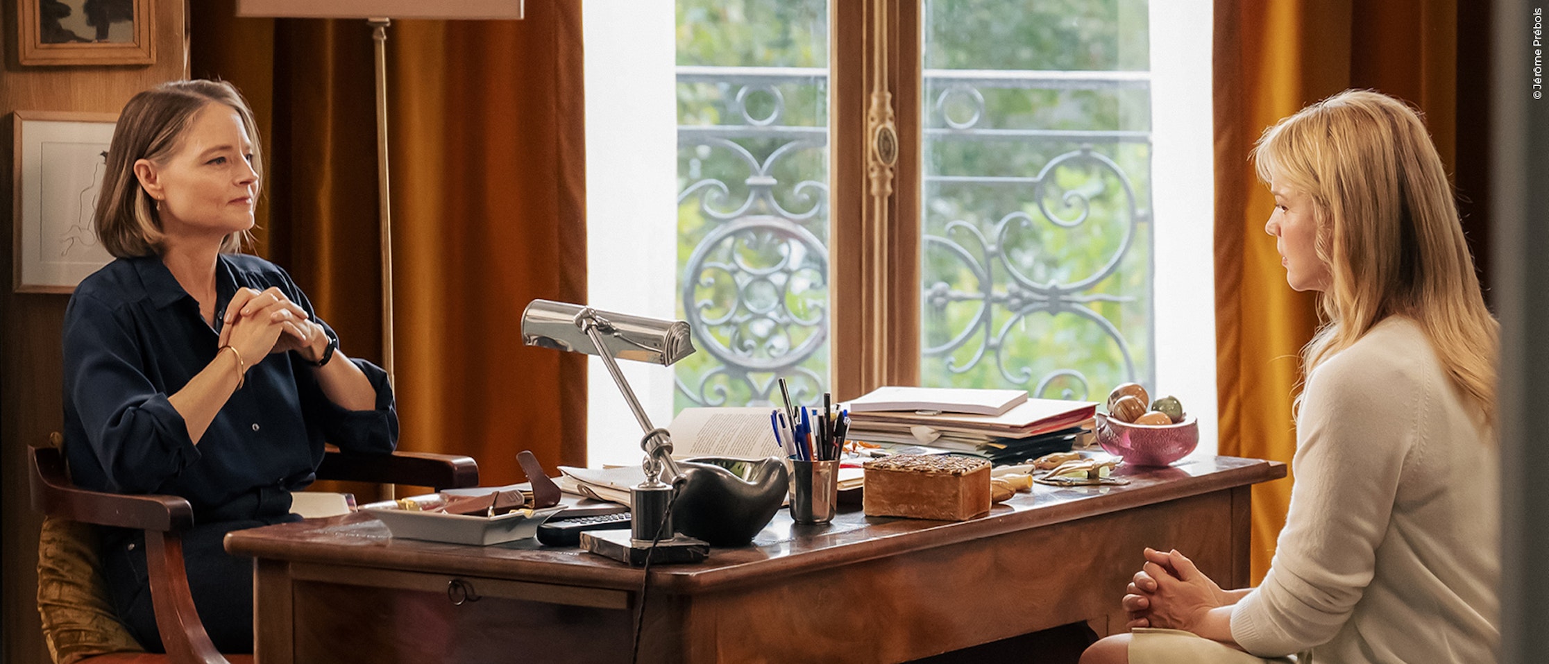 Zwei Frauen sitzen sich in einem hellen Büro mit großen Fenstern gegenüber. Die eine Frau sitzt hinter einem mit Büchern und Papieren vollgestopften Schreibtisch, während die andere ihr gegenüber sitzt und aufmerksam zuhört.