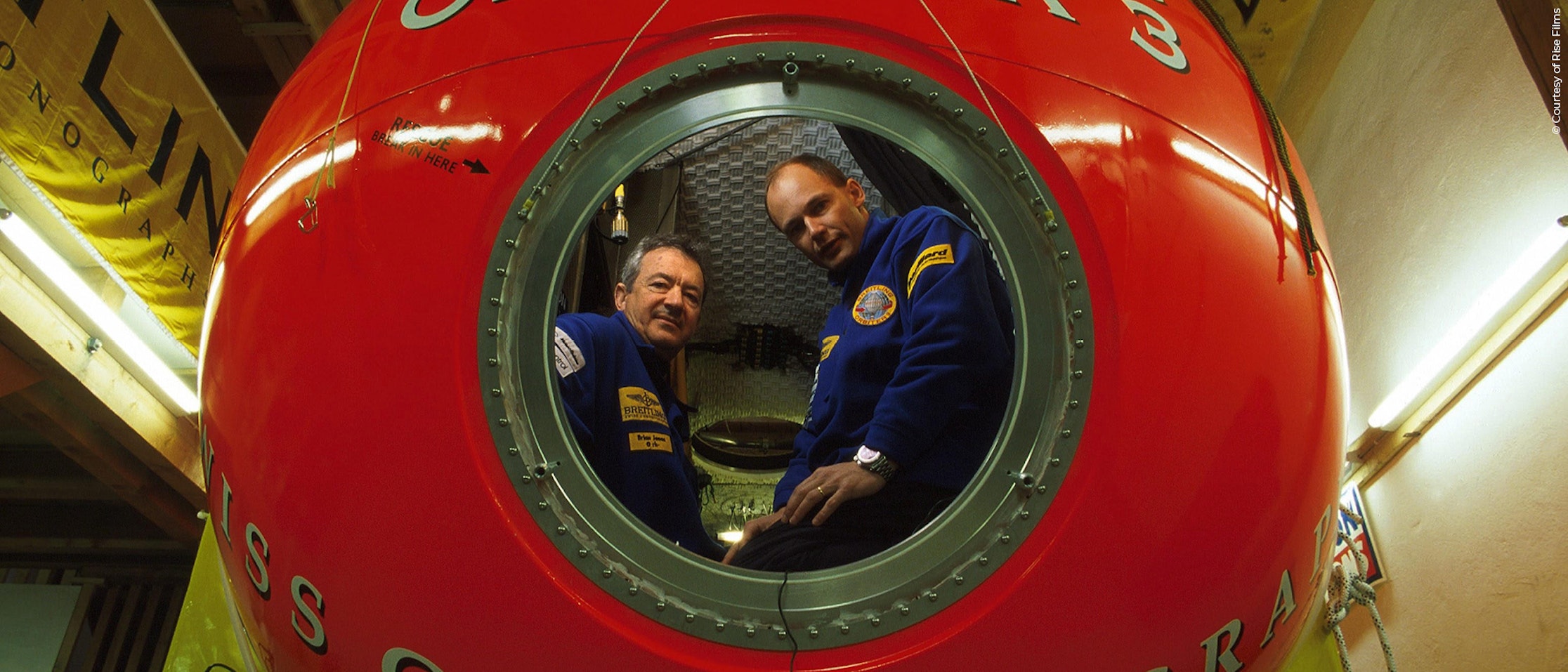 Zwei Männer in blauen Uniformen sitzen in einer leuchtend roten kugelförmigen Kapsel, die durch ein rundes Fenster zu sehen ist. Das Innere wirkt mechanisch, mit sichtbaren Zifferblättern und Instrumenten um sie herum.