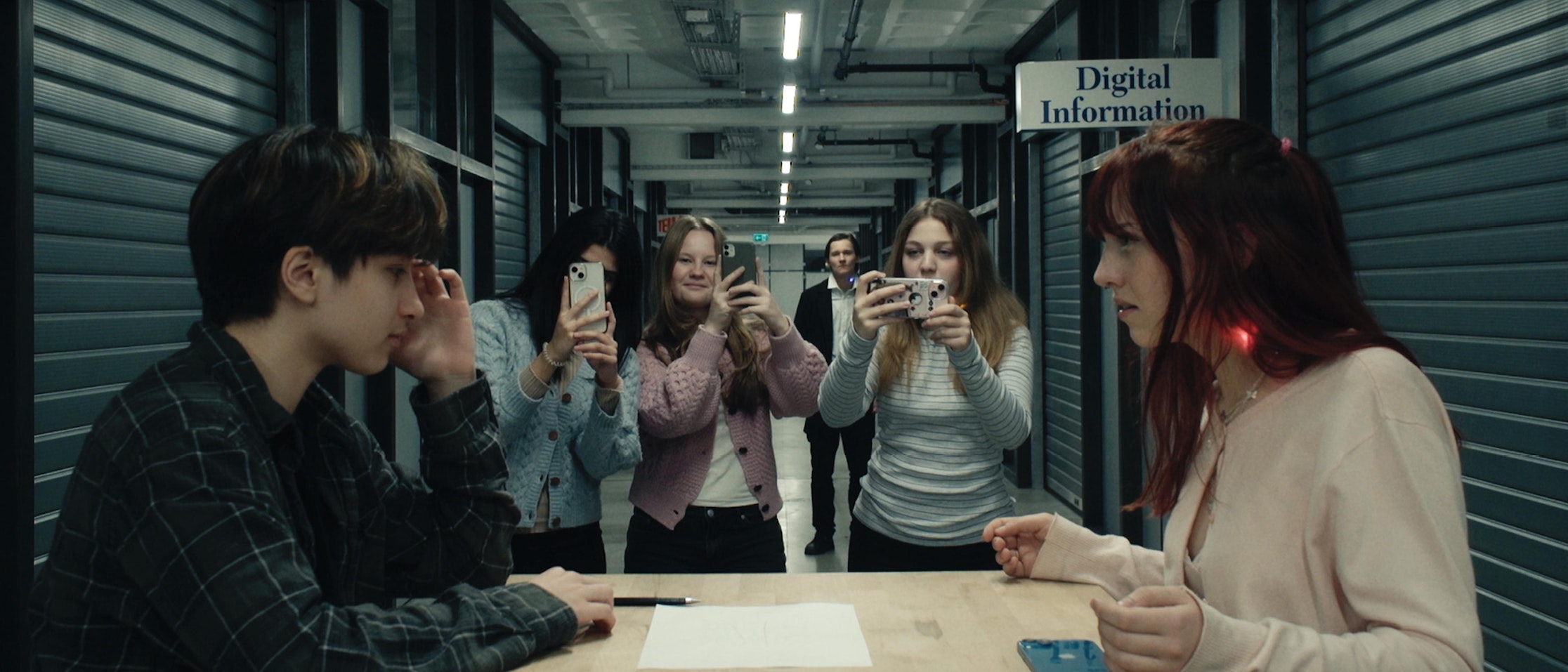 Zwei junge Leute sitzen sich an einem Tisch in einem Flur gegenüber, während vier andere hinter ihnen stehen und mit ihren Handys Fotos oder Videos aufnehmen. Darüber hängt ein Schild mit digitalen Informationen. Die Umgebung wirkt angespannt und modern.