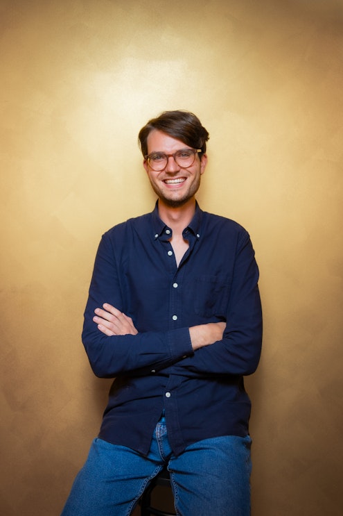 Eine Person mit kurzen dunklen Haaren und Brille, die ein marineblaues Button-up-Hemd und blaue Jeans trägt, sitzt auf einem Hocker und lächelt mit verschränkten Armen vor einem warmen gelben Hintergrund.