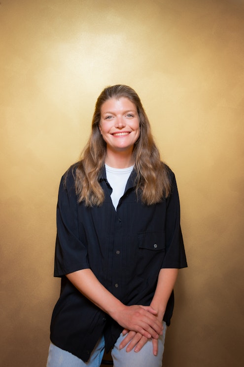 Eine Frau mit langen braunen Haaren sitzt lächelnd vor einem goldenen Hintergrund und trägt ein schwarzes Button-up-Hemd über einem weißen Top und hellblaue Jeans. Ihre Hände sind auf dem Schoß gefaltet.