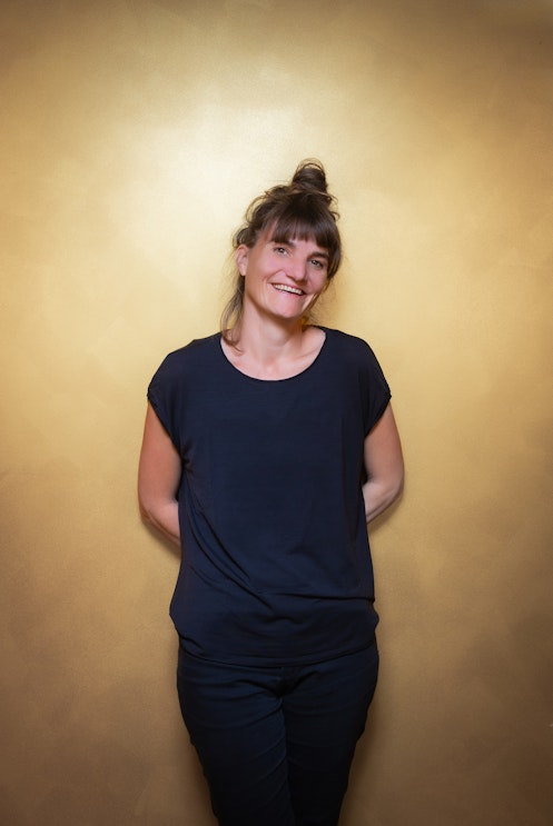 Eine Frau mit braunen Haaren in einem Dutt, die ein dunkelblaues T-Shirt und eine Hose trägt, steht lächelnd mit den Händen auf dem Rücken vor einer gelben Wand.