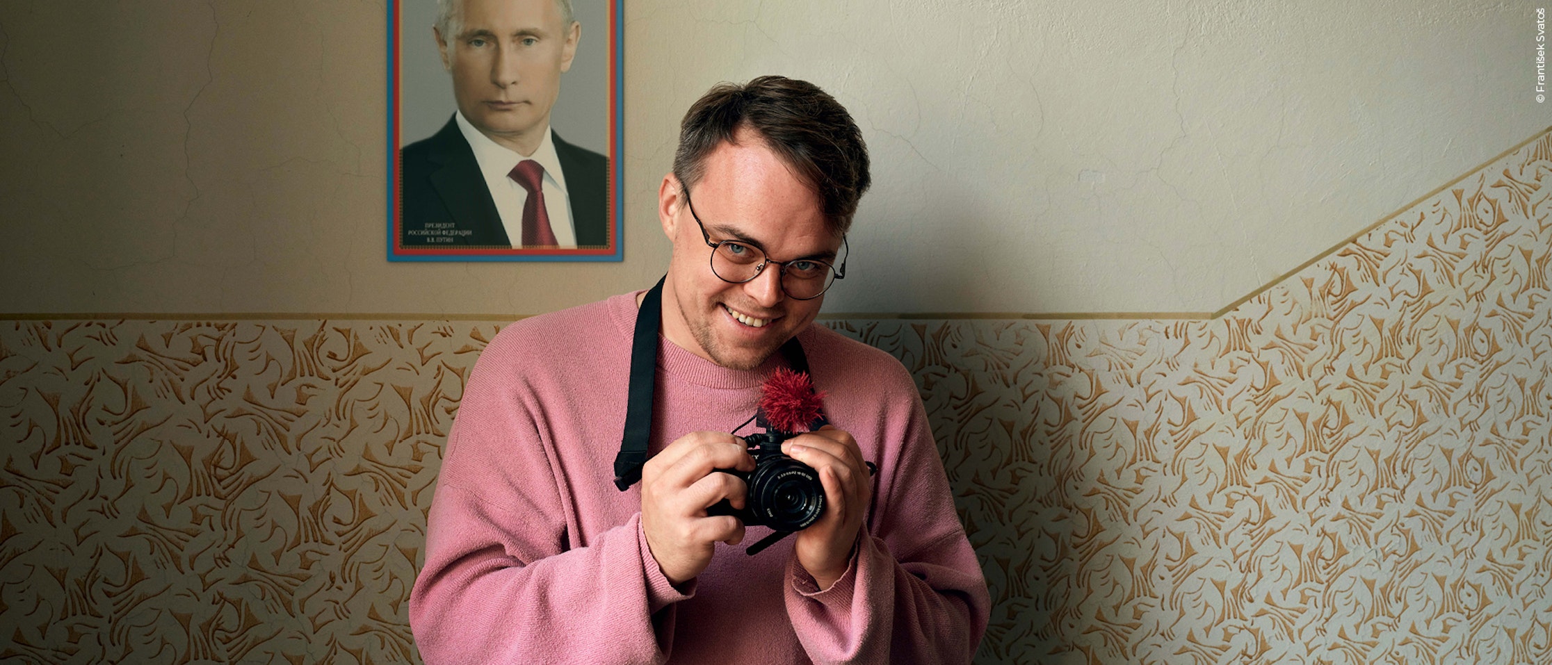 Ein Mann in einem rosa Pullover und mit Brille hält lächelnd eine Kamera in die Höhe. Hinter ihm hängt ein gerahmtes Porträt von Wladimir Putin an einer Wand mit gemusterter Tapete.