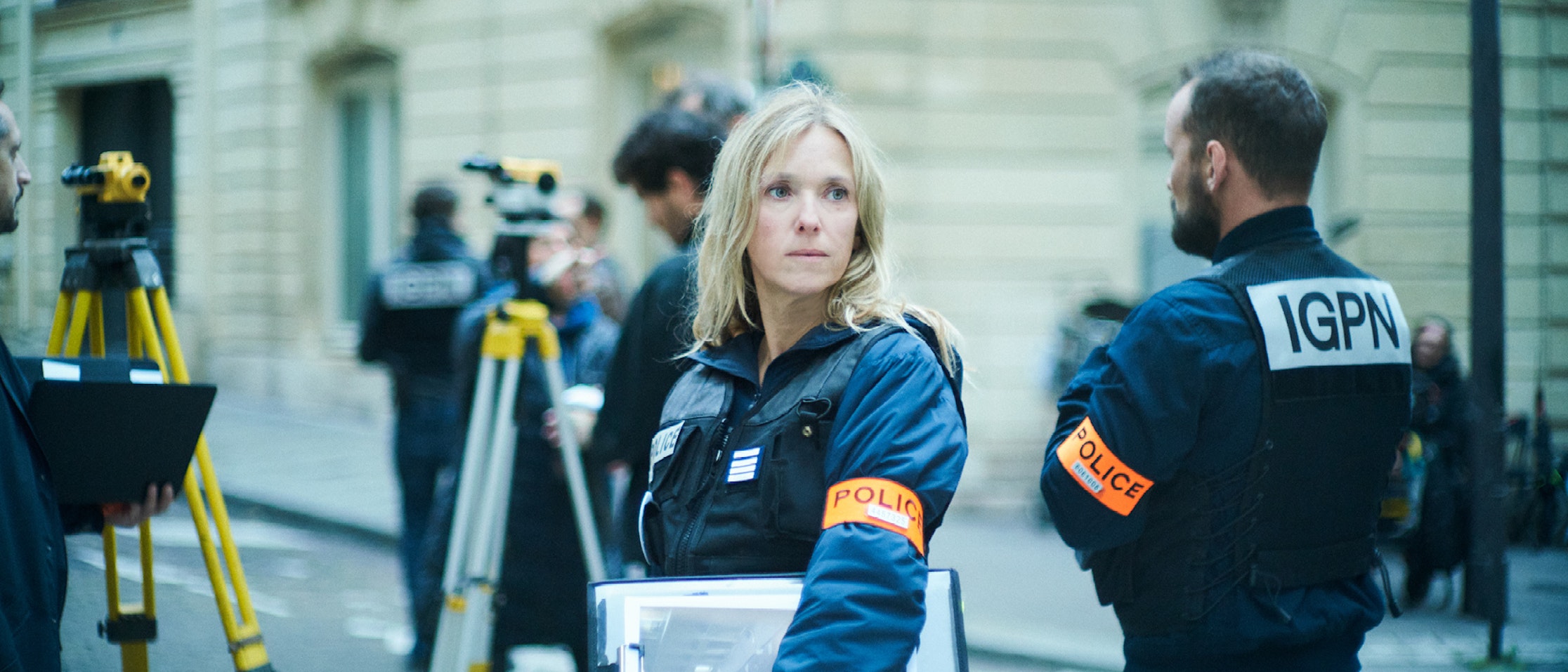 Eine weibliche Polizeibeamtin in Uniform mit einer orangefarbenen POLIZEI-Armbinde steht im Freien und hält Dokumente in der Hand, umgeben von Kollegen und Vermessungsgeräten auf einer städtischen Straße.
