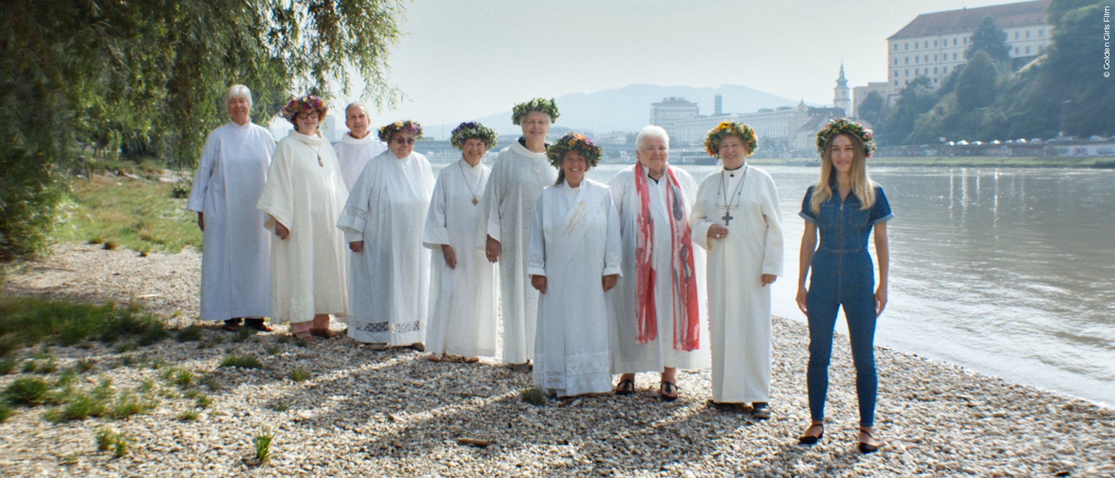 Eine Gruppe von neun Frauen in weißen Gewändern und Blumenkronen steht an einem felsigen Flussufer, wobei eine Frau in blauer Kleidung und mit einer Blumenkrone im Vordergrund steht. Im Hintergrund sind Bäume, Wasser und Gebäude zu sehen.