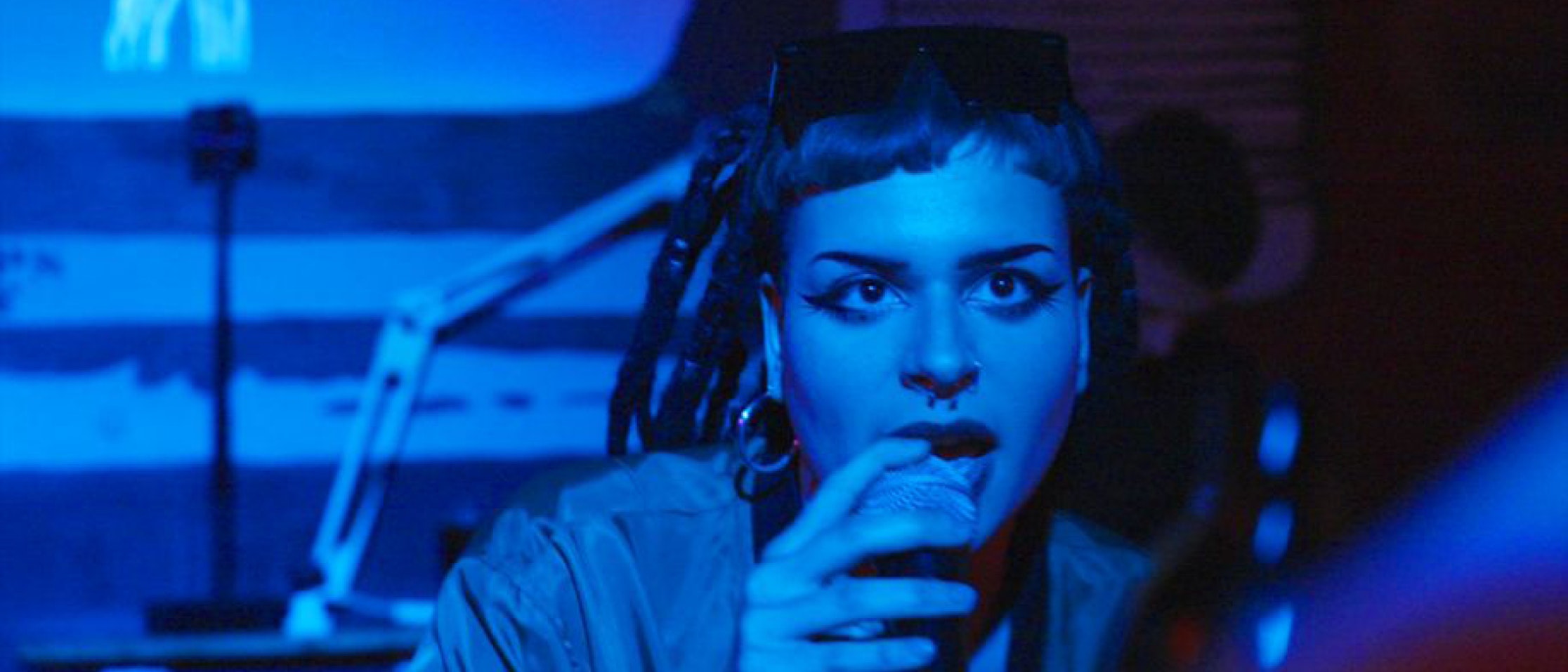 Eine Person mit kurzem Haar, auffälligem Eyeliner und einem Nasenring hält ein Mikrofon, während sie singt oder spricht. Die Szene ist in blaues Licht getaucht, mit einem verschwommenen Hintergrund, der eine Schreibtischlampe und abstrakte Muster zeigt.