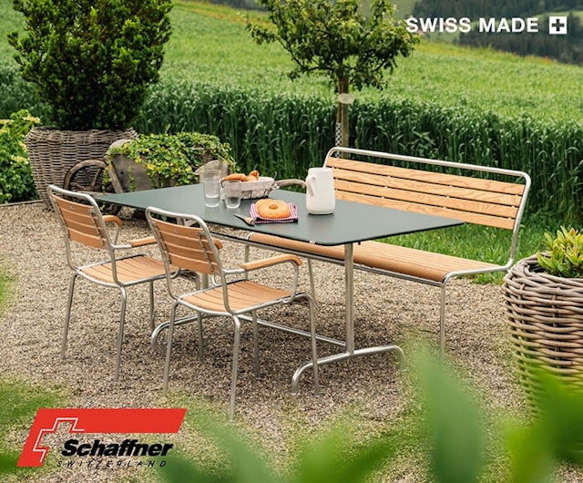 Essgruppe für den Außenbereich mit einem rechteckigen Tisch, zwei Stühlen und einer Bank mit Holzlatten und Metallrahmen. Der Tisch ist mit Frühstücksprodukten gedeckt, umgeben von Grünzeug und großen dekorativen Pflanzgefäßen. Logos von Schaffner Switzerland und Swiss Made.