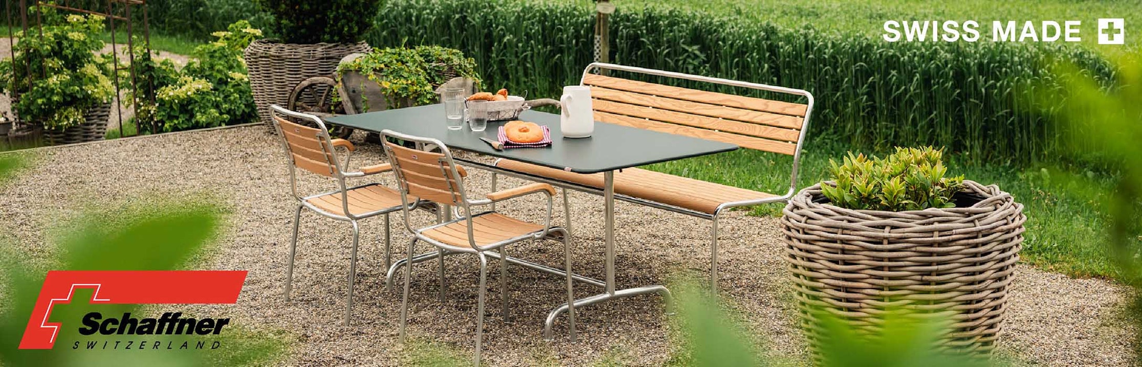 Esstisch im Freien mit Holz- und Metallstühlen auf einer Kiesterrasse, bestückt mit Tellern, Tassen und einer Kanne; umgeben von grünen Pflanzen und Feldern. Die Logos "Schaffner Switzerland" und "Swiss Made" erscheinen im Bild.