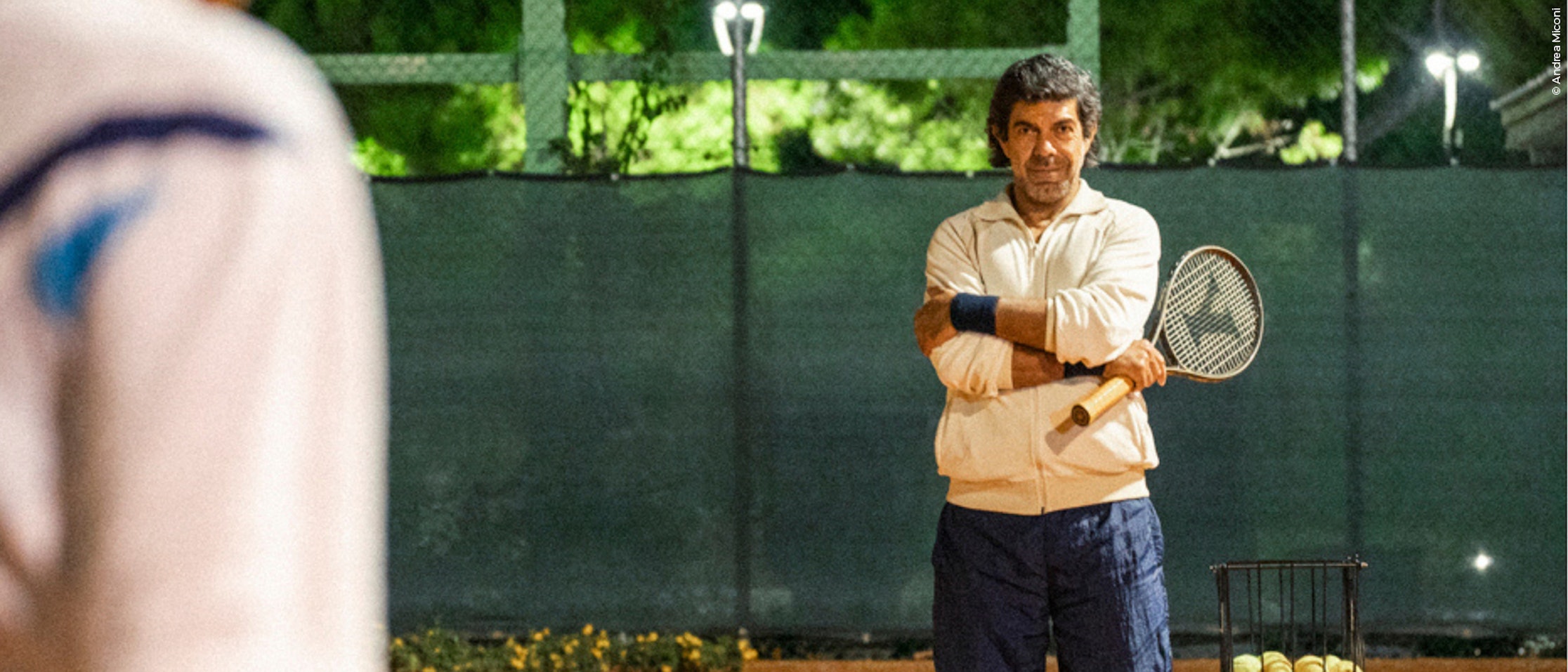 Ein Tennistrainer steht nachts auf einem Platz, die Arme verschränkt, einen Schläger in der Hand und einen Spieler vor sich. Ein Korb mit Tennisbällen steht in der Nähe, und helle Lichter erhellen die Szene.