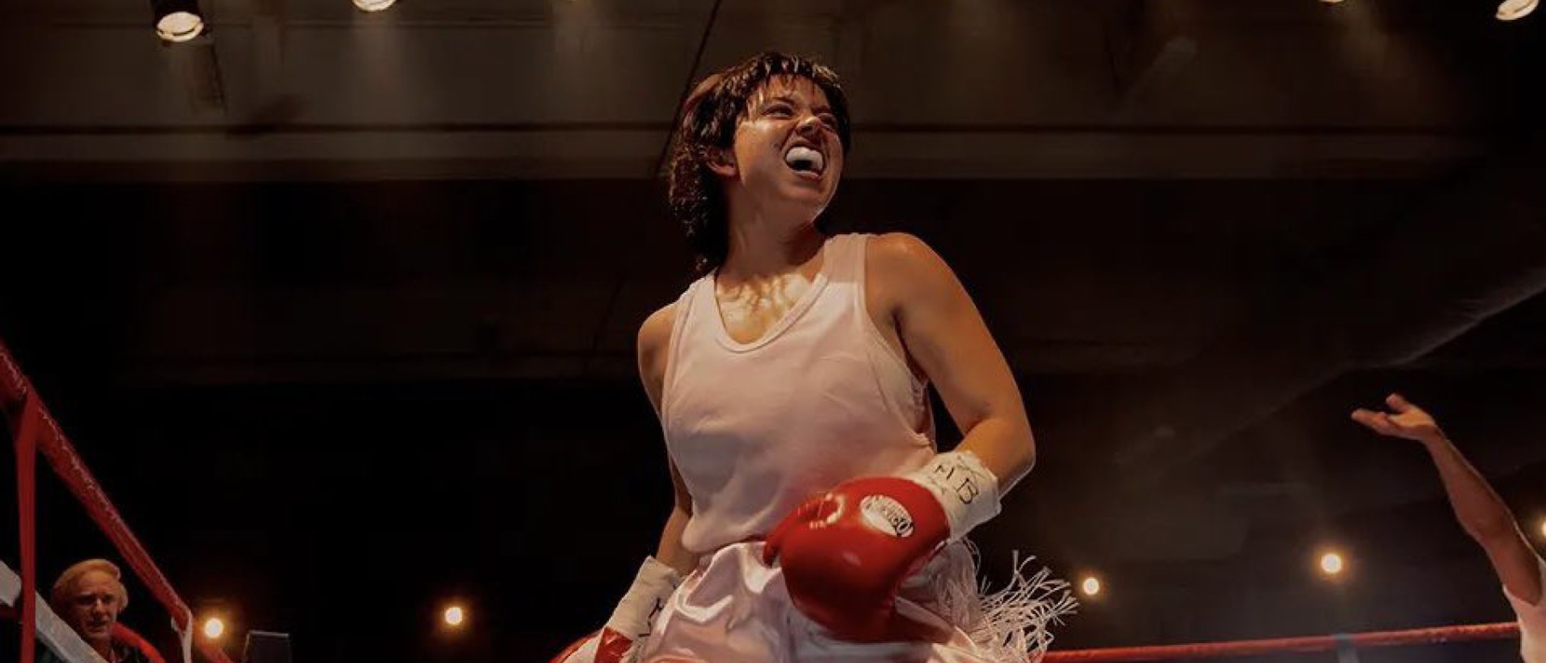 Ein Boxer mit roten Handschuhen und weißem Tank-Top steht in einem Boxring, blickt nach oben und schreit triumphierend, mit Schweiß im Gesicht und hellen Lichtern, die die Szene beleuchten.