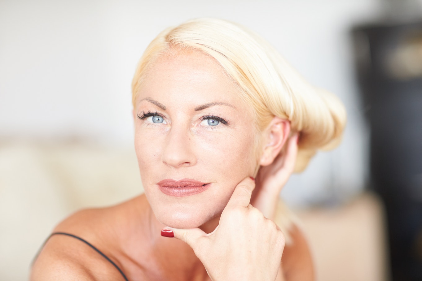 Eine Frau mit platinblondem Haar, das zu einer Hochsteckfrisur frisiert ist, blickt direkt in die Kamera und stützt ihr Kinn auf ihre Hand. Sie hat blaue Augen, ist leicht geschminkt und trägt ein Oberteil mit dünnen Trägern. Der Hintergrund ist sanft verschwommen.