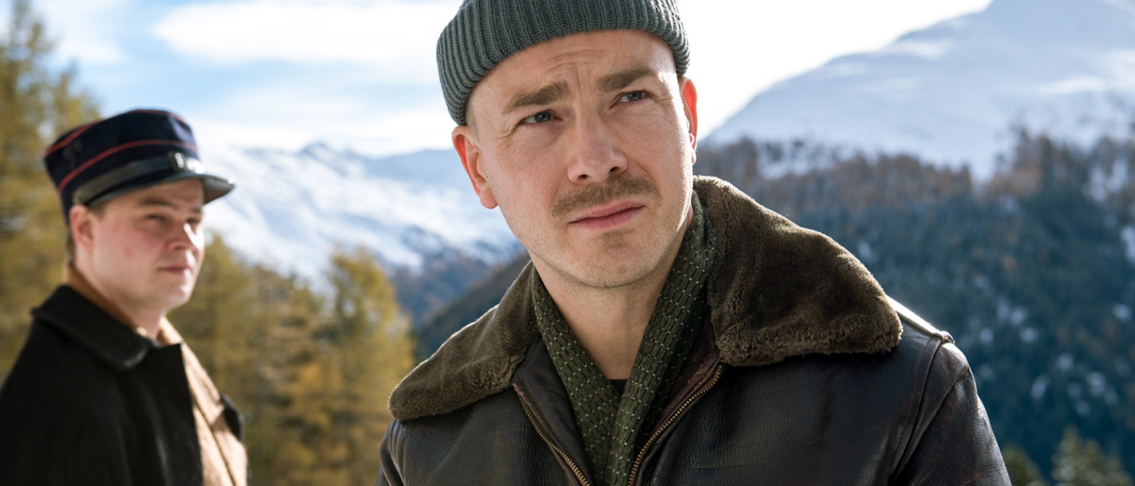 Zwei Männer stehen im Freien in einer verschneiten Berglandschaft. Der Mann im Vordergrund trägt eine grüne Strickmütze, eine braune Jacke und einen Schal, während der Mann im Hintergrund einen einheitlichen Hut und Mantel trägt. Sie sind von Kiefern und schneebedeckten Gipfeln umgeben.