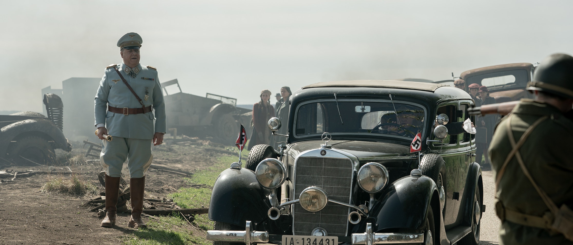 Ein Mann in einer deutschen Militäruniform aus der Zeit des Zweiten Weltkriegs steht neben einem schwarzen Oldtimer mit Nazi-Fahnen, während im Hintergrund Soldaten und beschädigte Fahrzeuge in einer verrauchten, vom Krieg zerstörten Landschaft zu sehen sind.