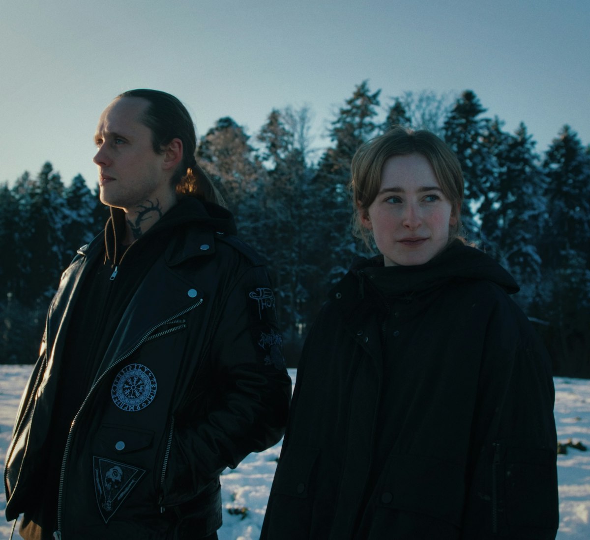 JONAS_ULRICH_WOLVES_STILL Zwei Personen in dunklen Jacken stehen im Freien auf einem verschneiten Feld mit einem Wald aus hohen Bäumen im Hintergrund. Die Sonne steht tief und wirft ein weiches Licht auf die Winterlandschaft.
