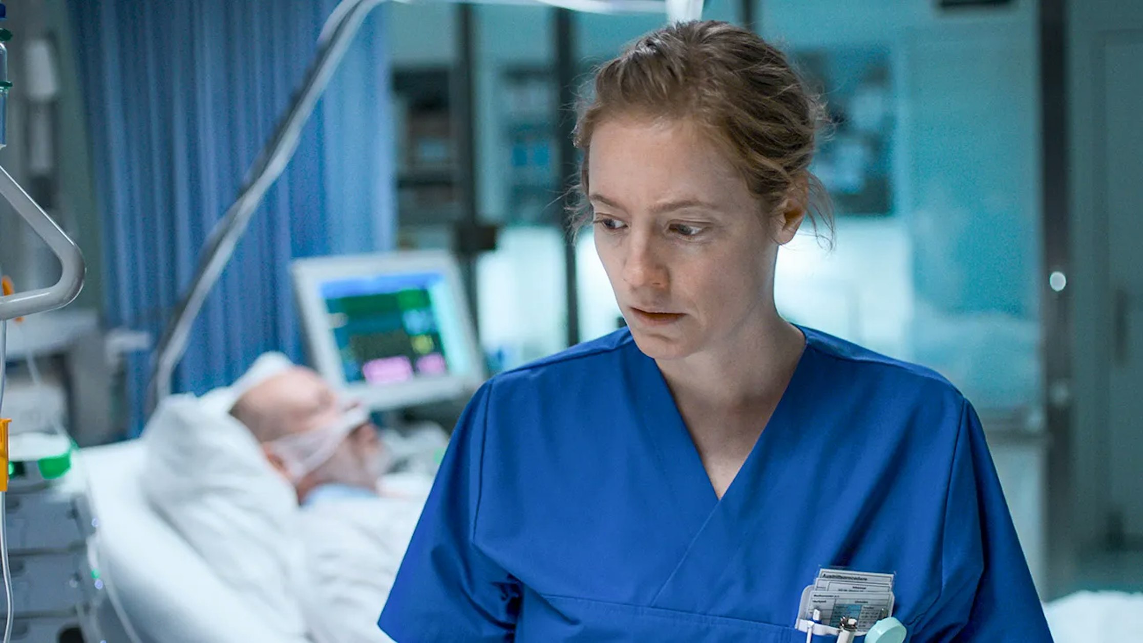 Eine Krankenschwester in blauem Kittel steht in einem Krankenhauszimmer und sieht besorgt aus. Im Hintergrund liegt ein Patient in einem Krankenhausbett, angeschlossen an Monitore und Sauerstoff, umgeben von medizinischen Geräten.