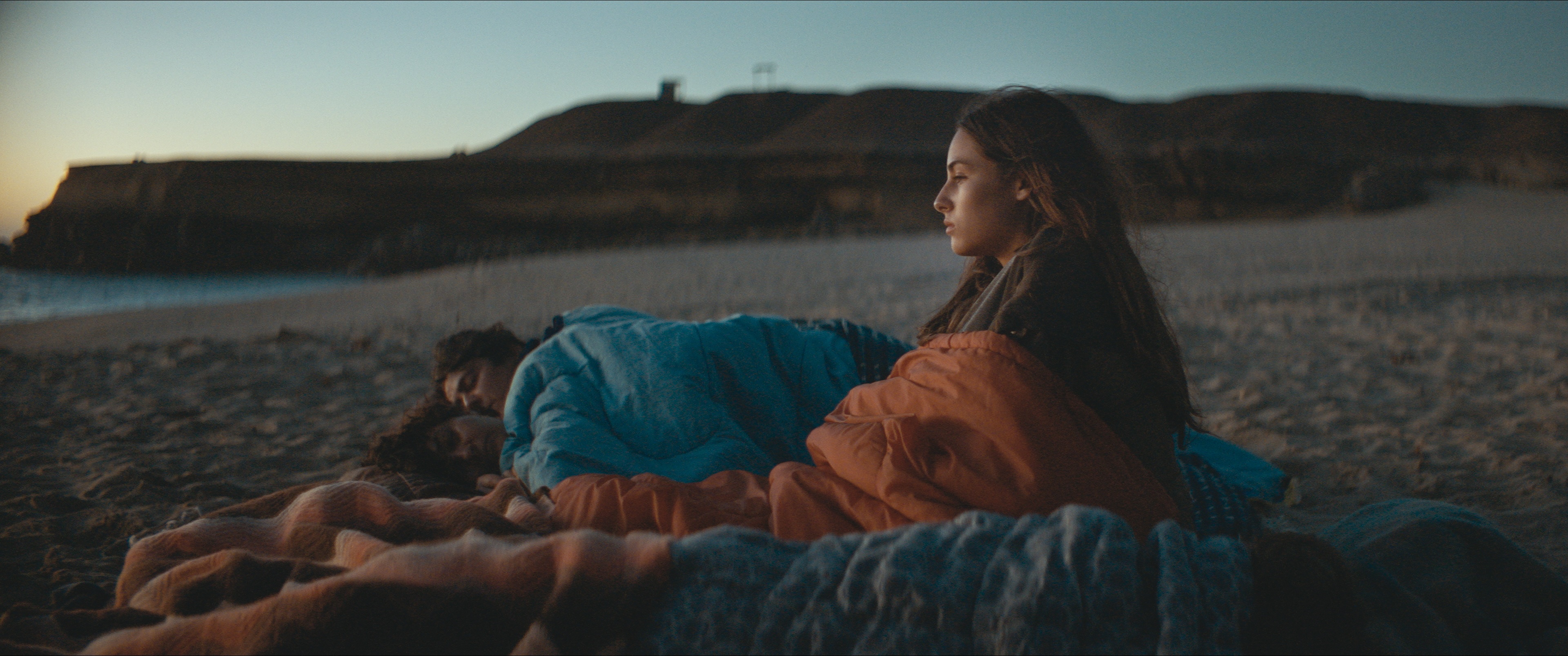Eine junge Frau sitzt in der Abenddämmerung an einem Strand, in eine Decke gehüllt, und blickt nachdenklich zum Horizont, während zwei Personen neben ihr unter Decken schlafen. Im Hintergrund sind felsige Klippen und das Meer zu sehen.