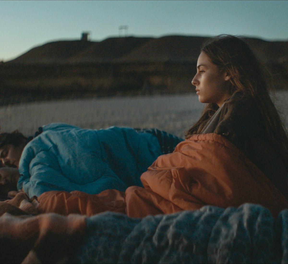 Cuerpo Celeste Film Still Eine junge Frau sitzt in der Abenddämmerung an einem Strand, in eine Decke gehüllt, und blickt nachdenklich zum Horizont, während zwei Personen neben ihr unter Decken schlafen. Im Hintergrund sind felsige Klippen und das Meer zu sehen.
