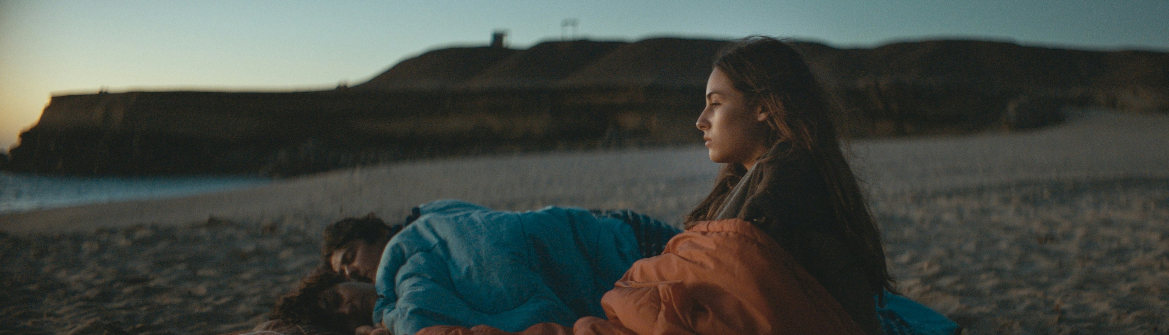 Cuerpo Celeste Film Still Eine junge Frau sitzt in der Abenddämmerung an einem Strand, in eine Decke gehüllt, und blickt nachdenklich zum Horizont, während zwei Personen neben ihr unter Decken schlafen. Im Hintergrund sind felsige Klippen und das Meer zu sehen.