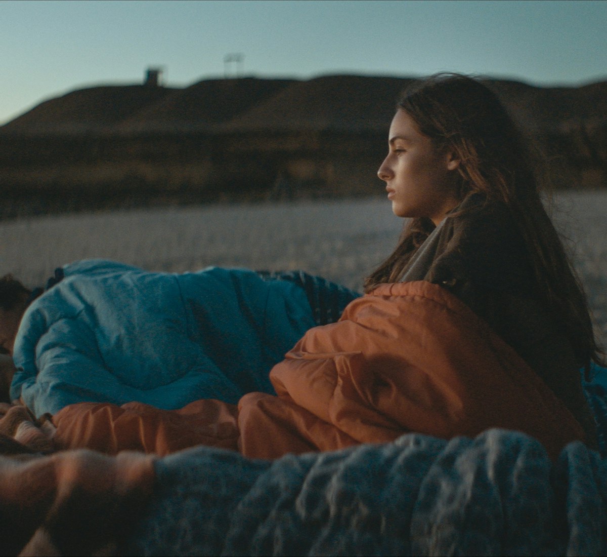 Film Still Cuerpo Celeste Eine junge Frau sitzt in der Abenddämmerung an einem Strand, in eine Decke gehüllt, und blickt nachdenklich zum Horizont, während zwei Personen neben ihr unter Decken schlafen. Im Hintergrund sind felsige Klippen und das Meer zu sehen.
