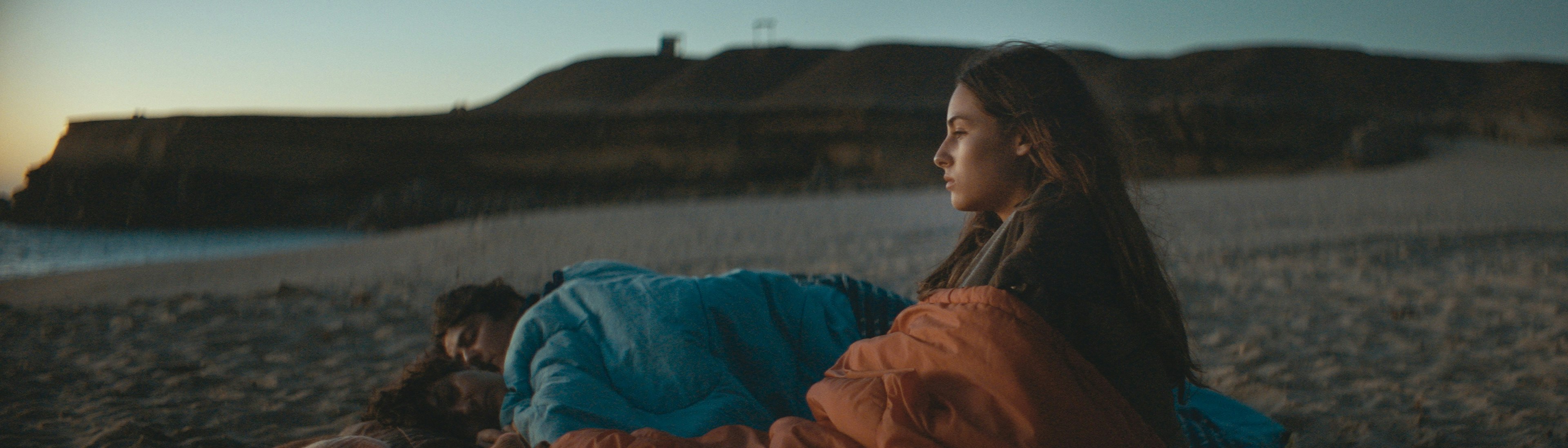 Film Still Cuerpo Celeste Eine junge Frau sitzt in der Abenddämmerung an einem Strand, in eine Decke gehüllt, und blickt nachdenklich zum Horizont, während zwei Personen neben ihr unter Decken schlafen. Im Hintergrund sind felsige Klippen und das Meer zu sehen.