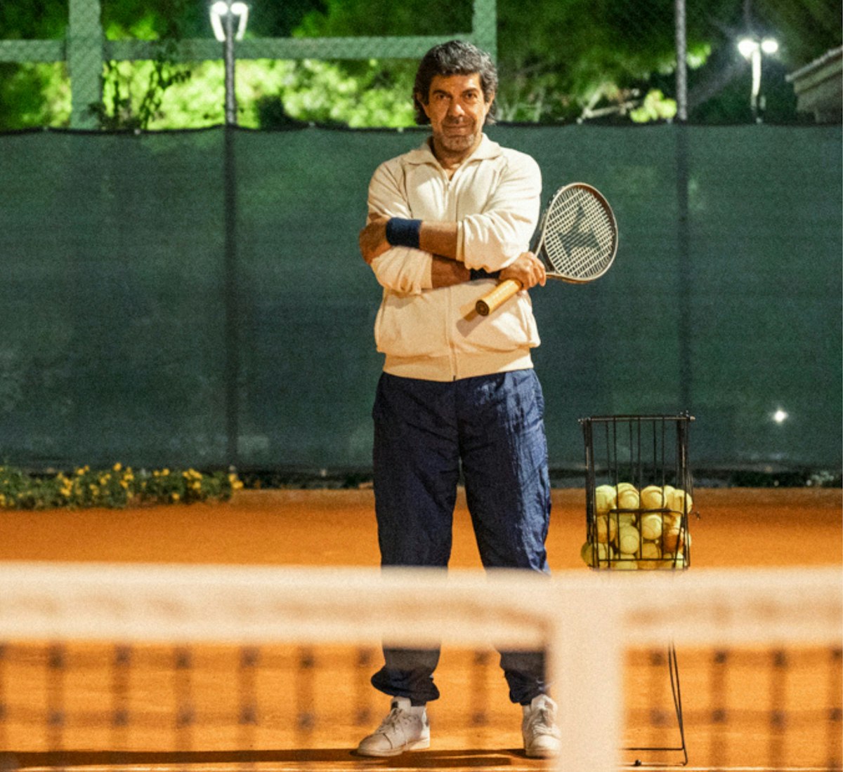 Il Maestro Film Still Ein Tennistrainer steht nachts auf einem Sandplatz, die Arme verschränkt, mit einem Schläger in der Hand, neben einem mit Tennisbällen gefüllten Korb. Die Szene wird von Oberlichtern beleuchtet, und im Vordergrund ist ein Spieler zu sehen.