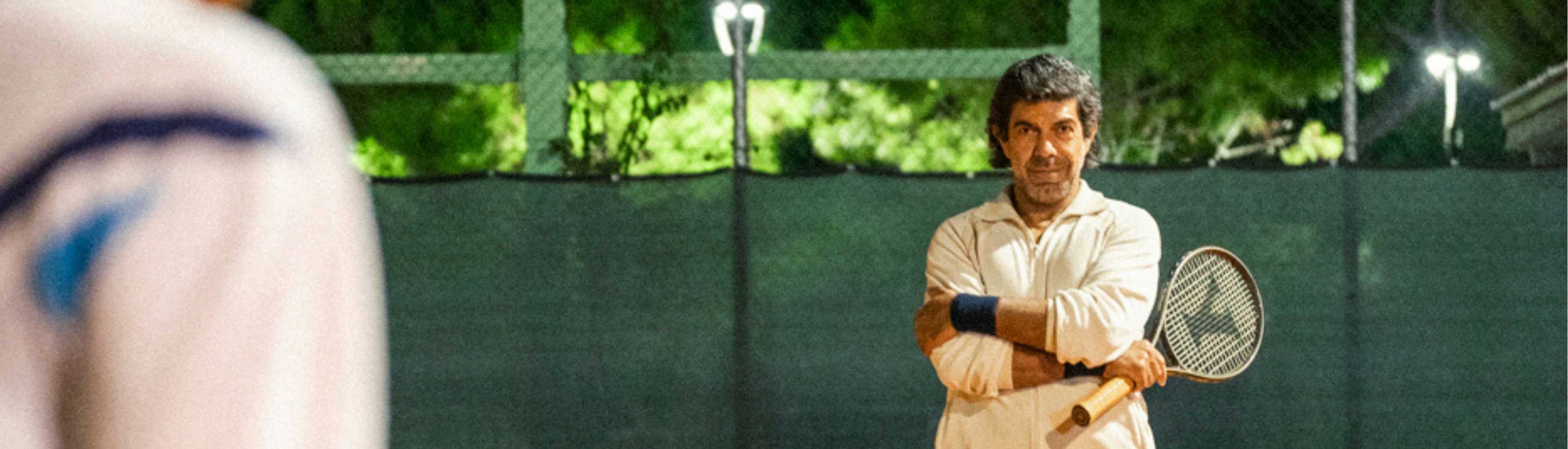 Il Maestro Film Still Ein Tennistrainer steht nachts auf einem Sandplatz, die Arme verschränkt, mit einem Schläger in der Hand, neben einem mit Tennisbällen gefüllten Korb. Die Szene wird von Oberlichtern beleuchtet, und im Vordergrund ist ein Spieler zu sehen.