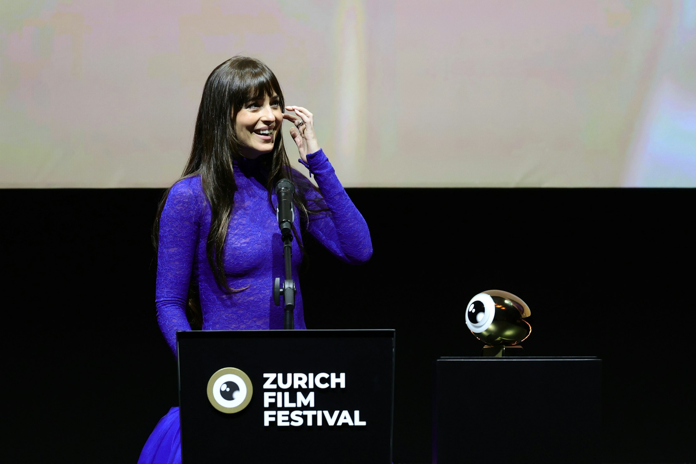 Eine Frau in einem langärmeligen, hellvioletten Kleid spricht an einem Podium mit der Aufschrift Zurich Film Festival, das neben einer goldenen, augenförmigen Trophäe auf einem schwarzen Ständer steht.