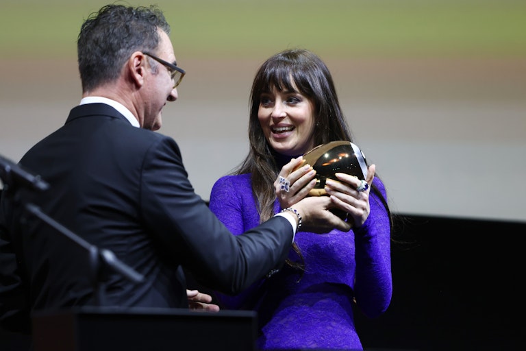 Eine Frau in einem lila Kleid lächelt, als sie während einer Preisverleihung eine goldene Trophäe von einem Mann im Anzug entgegennimmt.