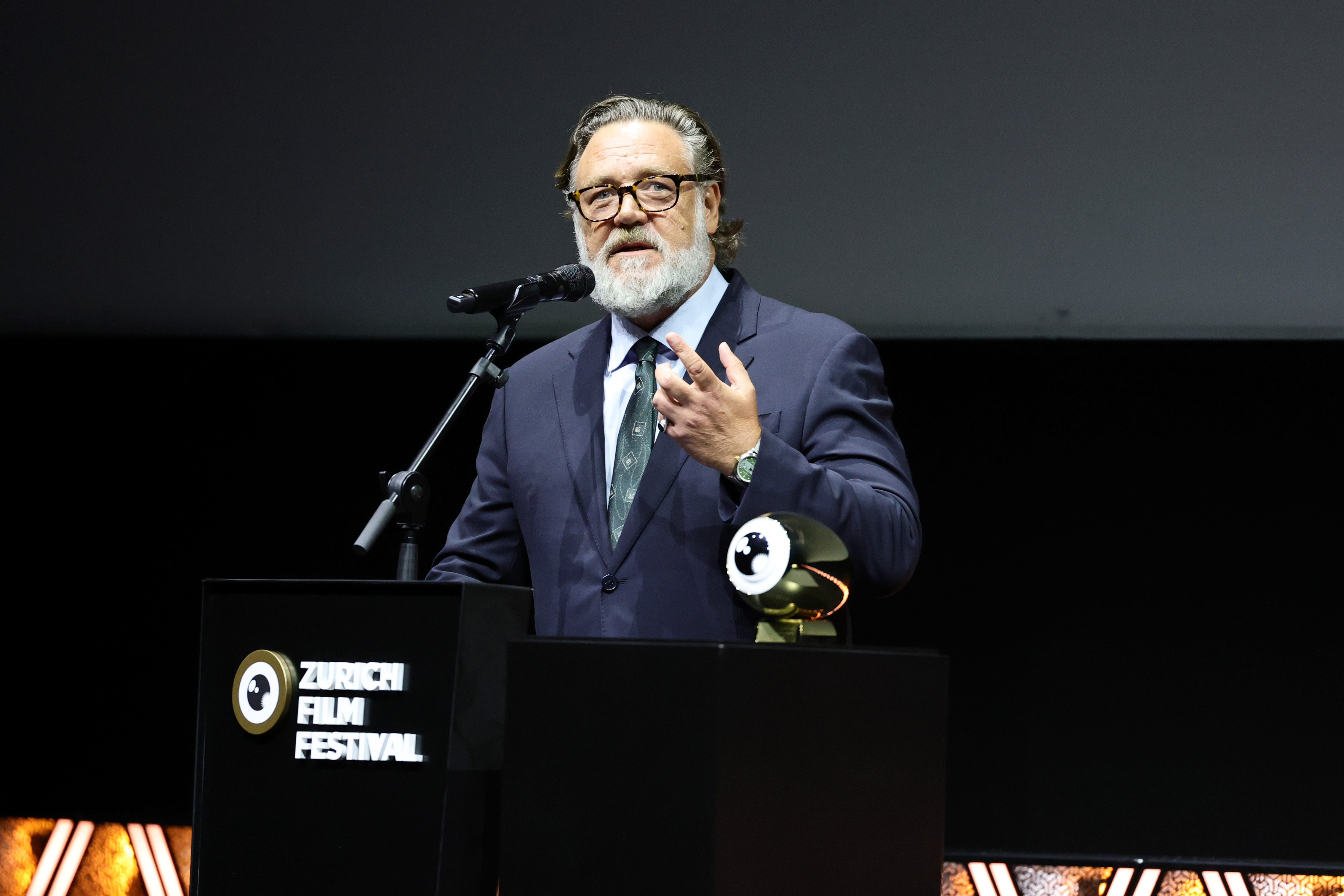 Ein Mann in Anzug und Krawatte spricht auf der Bühne des Zurich Film Festival auf einem Podium, neben ihm stehen ein Mikrofon und eine Trophäe.