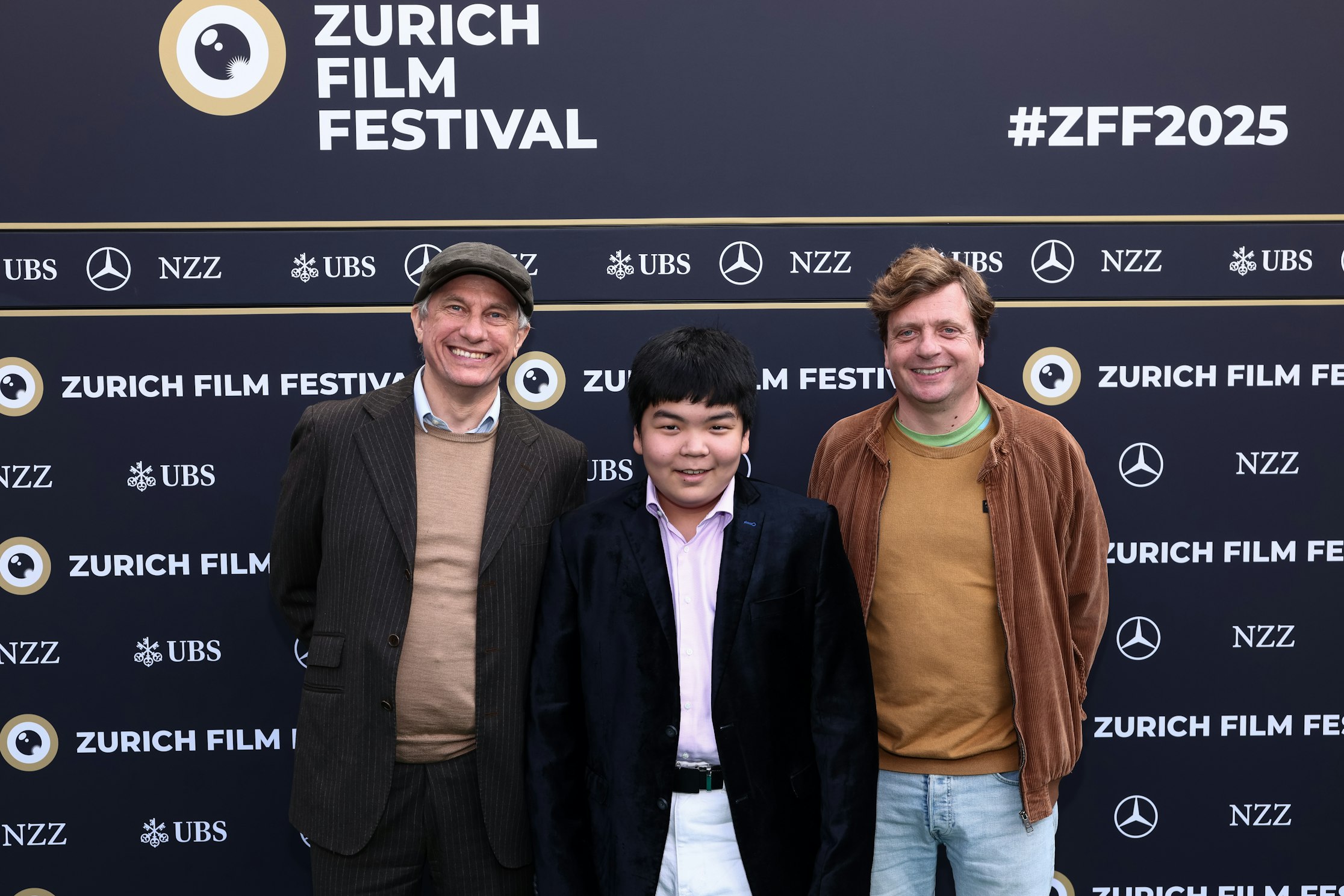 Drei Personen stehen lächelnd vor einer Kulisse des Zurich Film Festival, auf der Sponsorenlogos und "#ZFF2025" zu sehen sind. Sie sind halb-formal gekleidet, unter anderem mit Blazern und Pullovern.