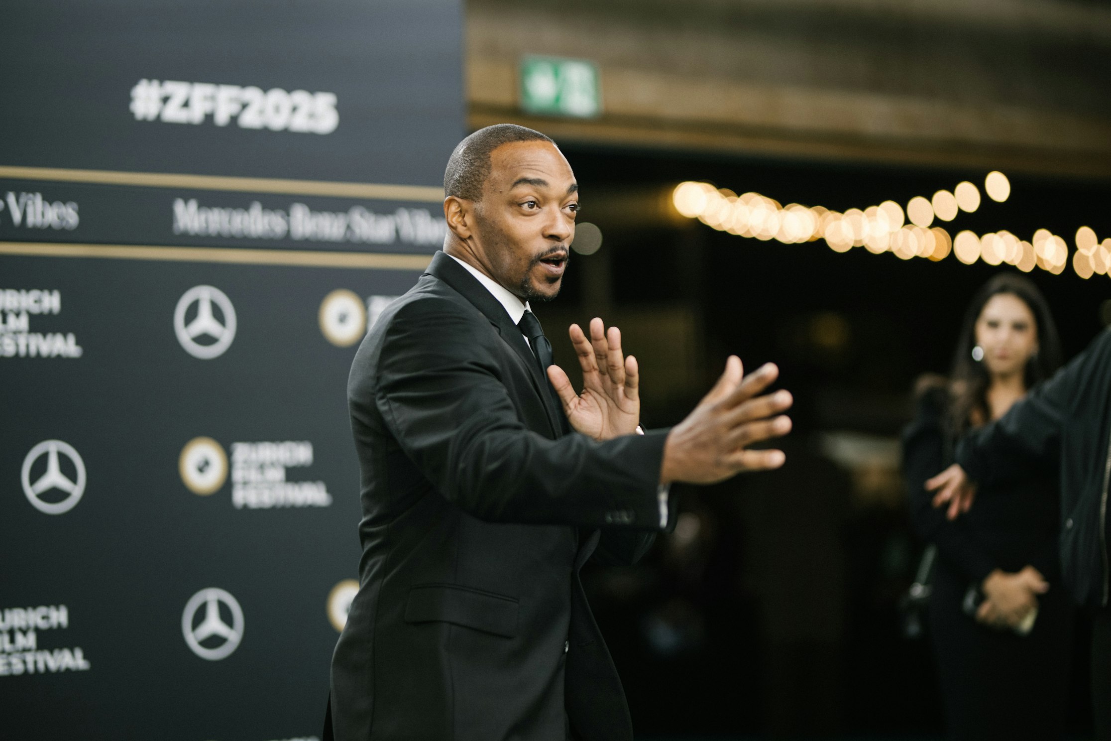 Ein Mann in einem schwarzen Anzug gestikuliert angeregt auf dem roten Teppich des Zurich Film Festival, mit Festival- und Sponsorenlogos und verschwommenen Lichtern im Hintergrund.