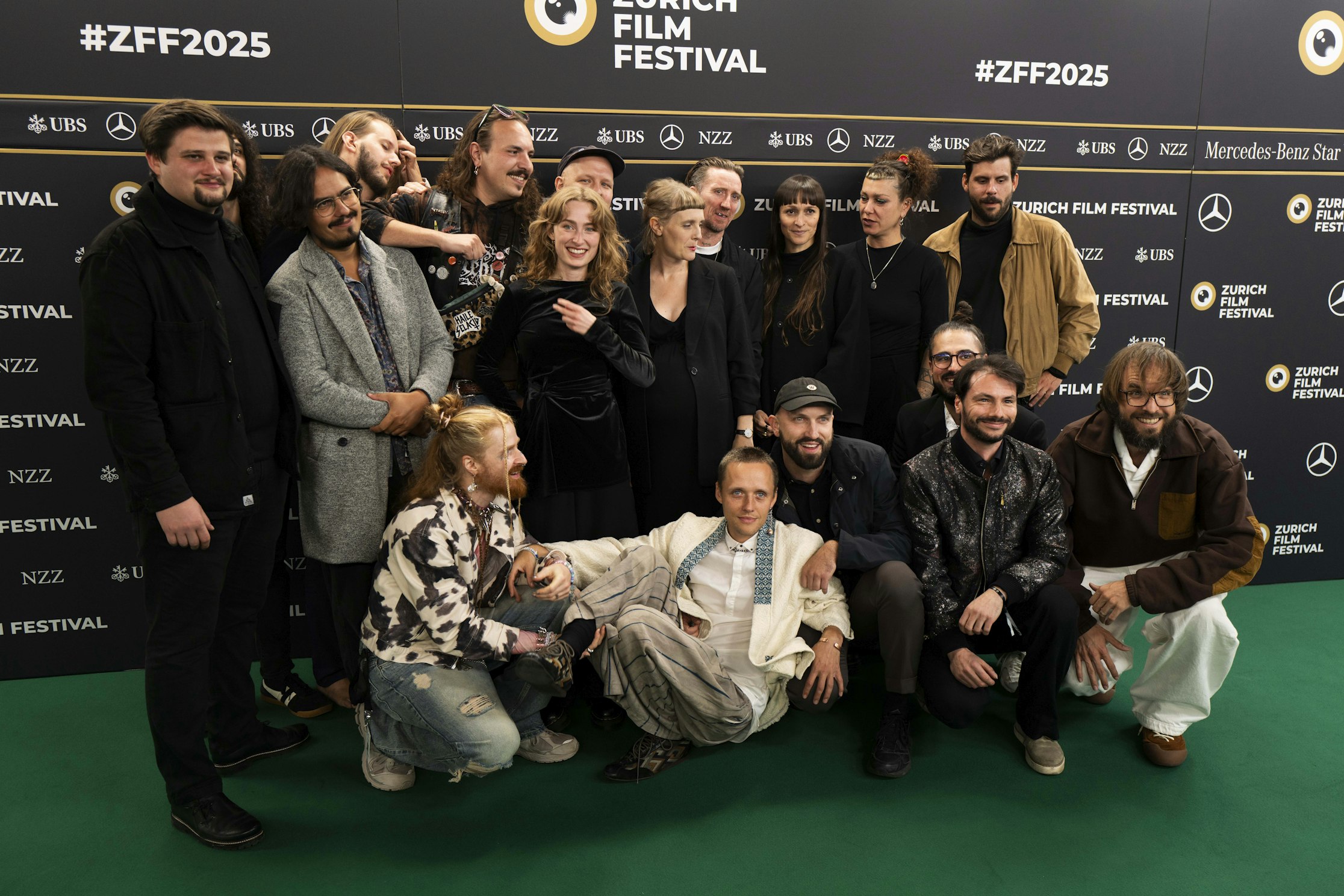 Eine grosse Gruppe von Menschen posiert gemeinsam lächelnd auf einem grünen Teppich vor einer Kulisse des Zurich Film Festival mit Logos und #ZFF2025. Einige stehen, andere knien in der ersten Reihe.