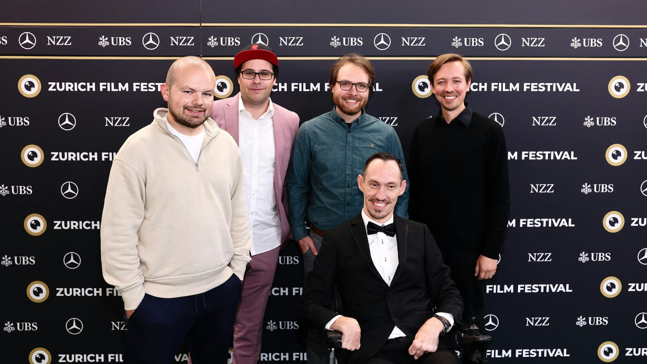 Fünf Männer posieren gemeinsam auf dem roten Teppich des Zurich Film Festival; einer sitzt in einem Rollstuhl und trägt einen Smoking, während die anderen hinter ihm stehen und vor einer Markenkulisse lächeln.